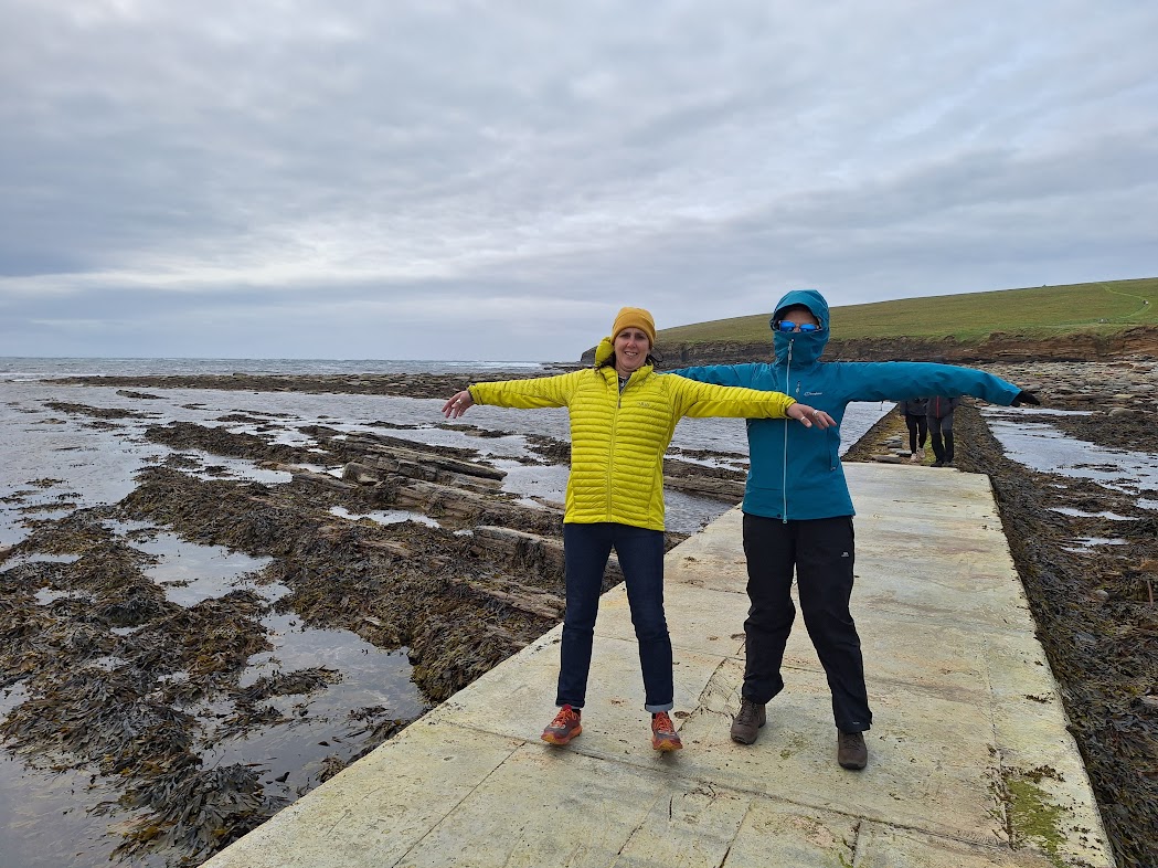 2 DMC members standing by the coast, arms outstretched, leaning into the wind